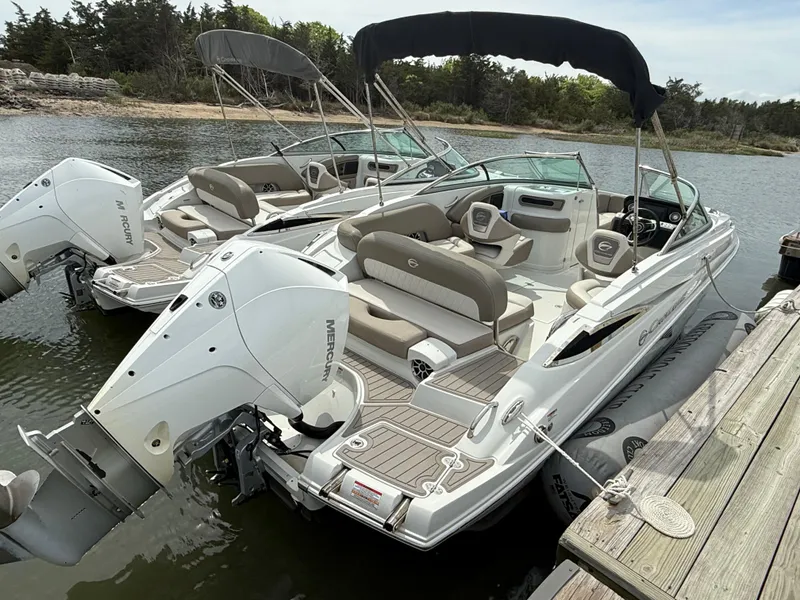 Slide: The Image of 2023 Crownline Eclipse E235 XS boat docked on a calm lake with dual outboard motors. - 2
