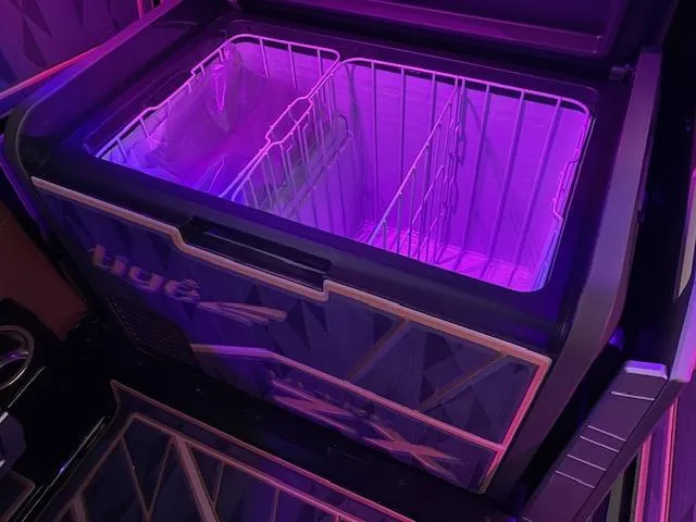 Slide: The Image of 2025 Tigé Ultré 25ZX boat cooler with purple lighting, open lid, and storage baskets. - 6