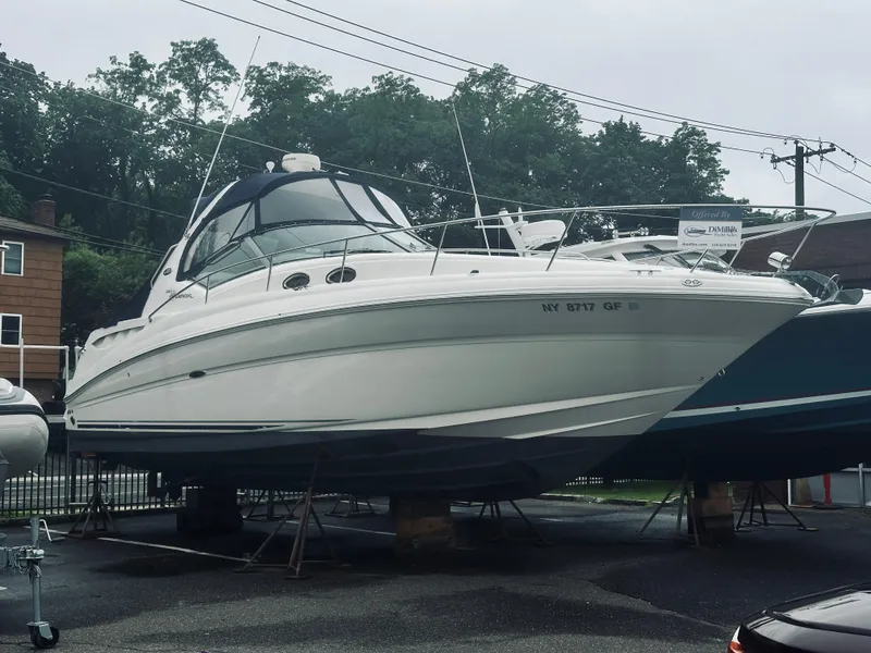 Slide: The Image of 2006 Sea Ray 320 Sundancer boat on stands, parked outdoors, surrounded by trees and buildings. - 0