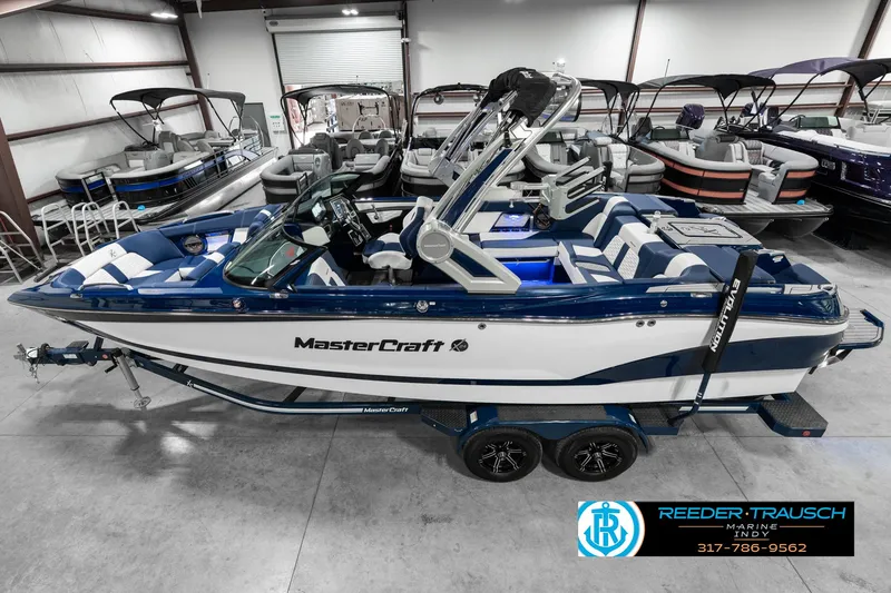 Slide: The Image of 2025 MasterCraft X24 boat in showroom, blue and white design, on trailer. - 1
