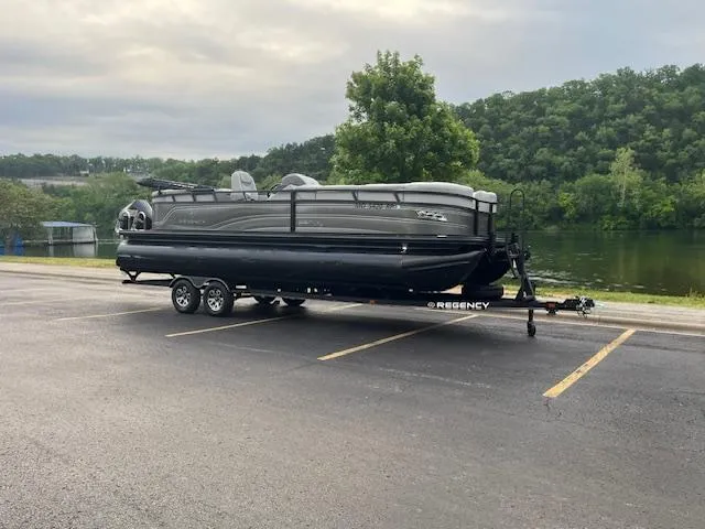 Slide: The Image of 2024 Regency 250LE3 pontoon boat on trailer, parked near a scenic river. - 0