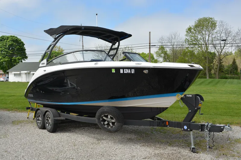 Slide: The Image of 2021 Yamaha Boats 252SD on trailer, black hull, parked on gravel, green lawn background. - 2
