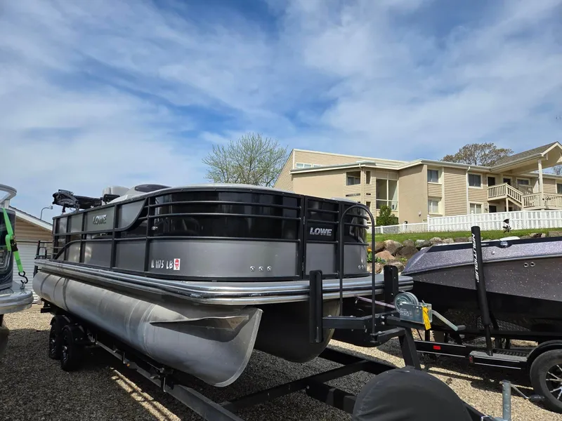 Slide: The Image of 2021 Lowe Retreat 230 High Performance pontoon boat on trailer, parked near residential building. - 5