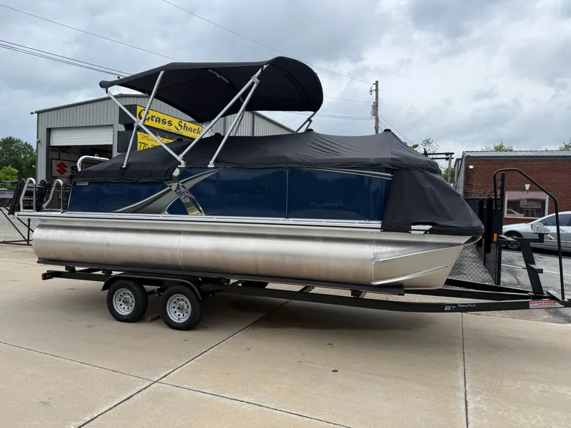 Slide: The Image of 2025 Avalon LSZ Quad Lounger Shift pontoon boat on trailer, parked outdoors. - 2