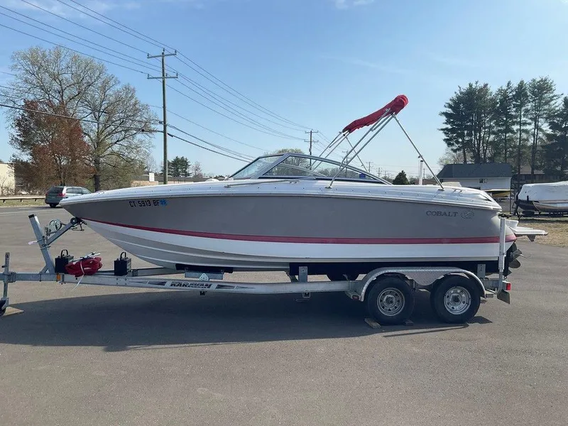 Slide: The Image of 2013 Cobalt 210 boat on trailer, parked outdoors, with trees and power lines in background. - 21