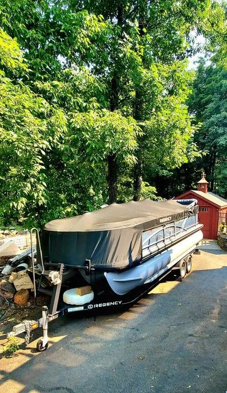 Slide: The Image of 2017 Regency 220 DL3 pontoon boat covered, parked on a driveway near trees and a shed. - 26