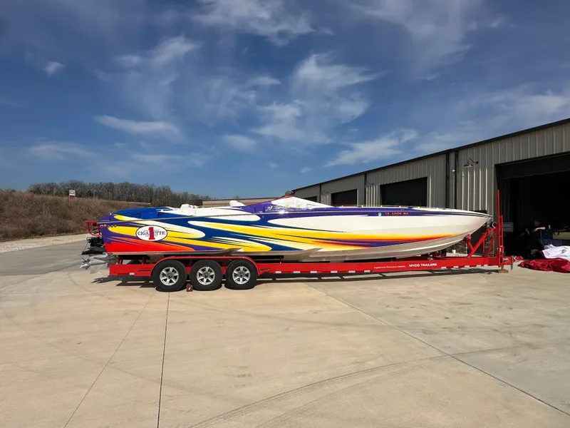 The Image of 2004 Cigarette 38 Top Gun boat on red trailer, vibrant graphics, parked outside a warehouse. - 1