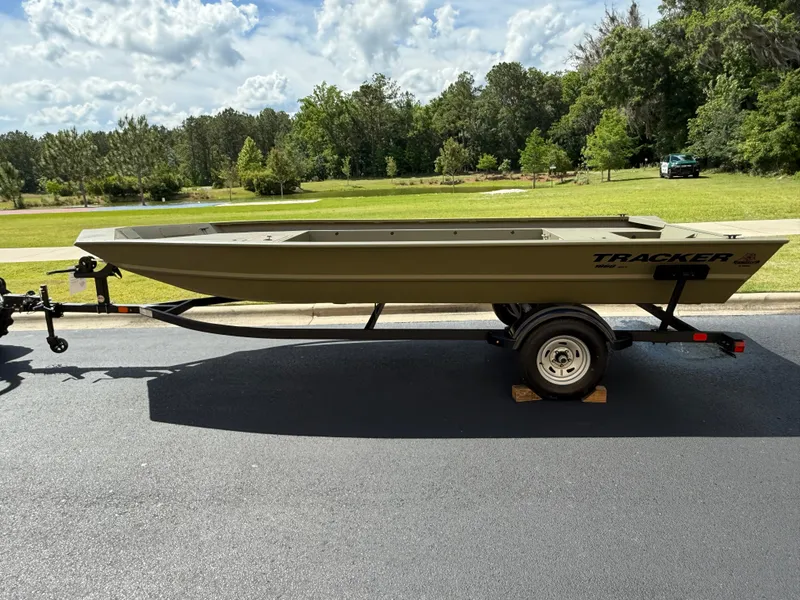 The Image of 2025 Tracker GRIZZLY 1860 Utility boat on trailer, parked on asphalt, with lush greenery background. - 0