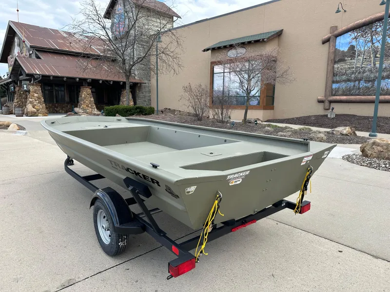Slide: The Image of 2025 Tracker GRIZZLY 1860 Utility boat on trailer, parked outdoors near rustic building. - 2