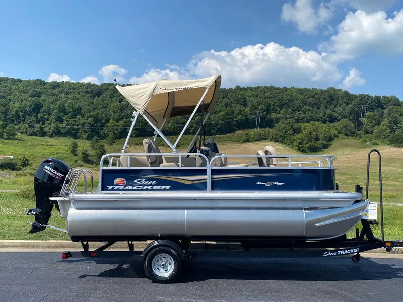 Slide: The Image of 2025 Sun Tracker Bass Buggy 16 XL Select pontoon boat on trailer, scenic background. - 2