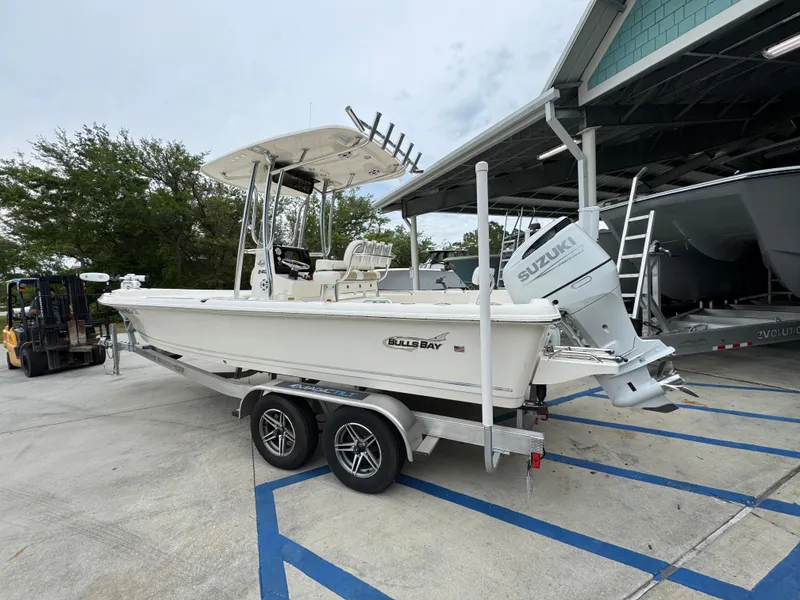 Slide: The Image of 2024 Bulls Bay 2400 boat on trailer with Suzuki outboard motor, parked near a building. - 18