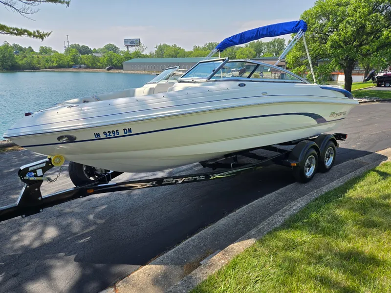 Slide: The Image of 2013 Bryant 210 STE boat on trailer by a lake, featuring blue canopy. - 8