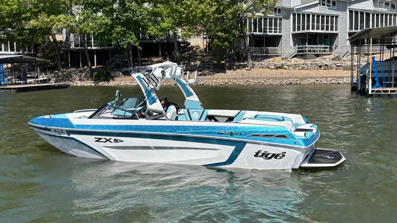 Slide: The Image of 2019 Tigé 25 ZXS boat on water, near lakeside homes, featuring sleek blue and white design. - 11