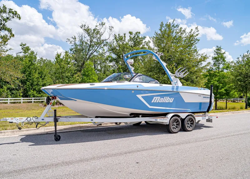 Slide: The Image of 2024 Malibu Wakesetter 24 MXZ boat on trailer, parked on road with trees in background. - 1