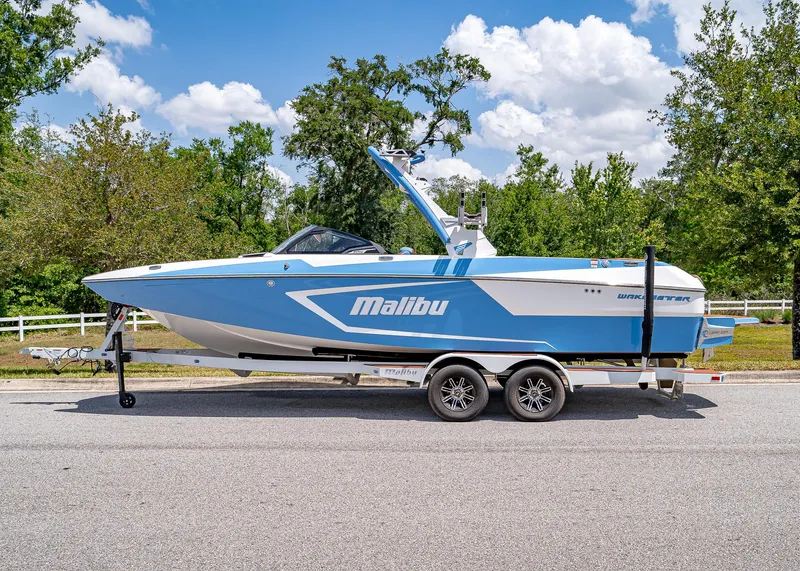 Slide: The Image of 2024 Malibu Wakesetter 24 MXZ boat on trailer, parked on road, surrounded by trees. - 2