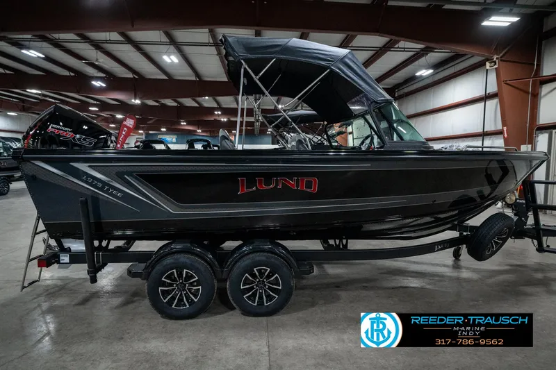 Slide: The Image of 2025 Lund 1975 Tyee boat in showroom, black exterior, dual-axle trailer, Reeder-Trausch Marine logo. - 6