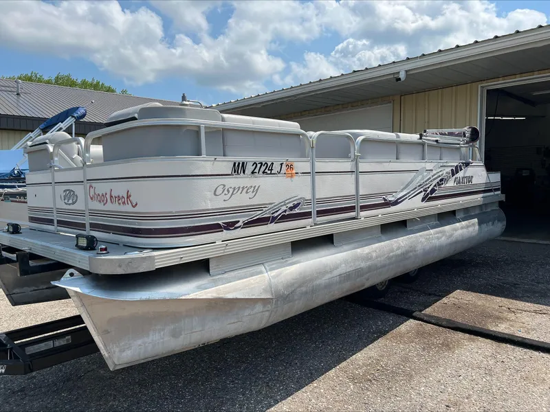 Slide: The Image of 2003 Manitou Osprey pontoon boat parked outside a garage under a cloudy sky. - 1