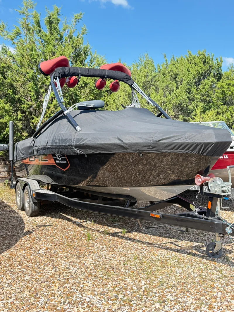 Slide: The Image of 2012 Epic 23V boat on trailer, covered, surrounded by trees and gravel. - 1