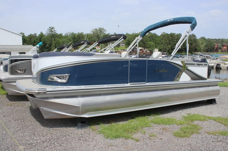 Slide: The Image of 2024 Avalon LSZ Quad Lounger pontoon boat docked on gravel near a marina. - 0