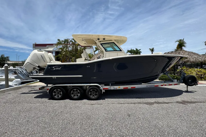 The Image of 2025 Scout 305 LXF boat on trailer, parked near marina under clear sky. - 0