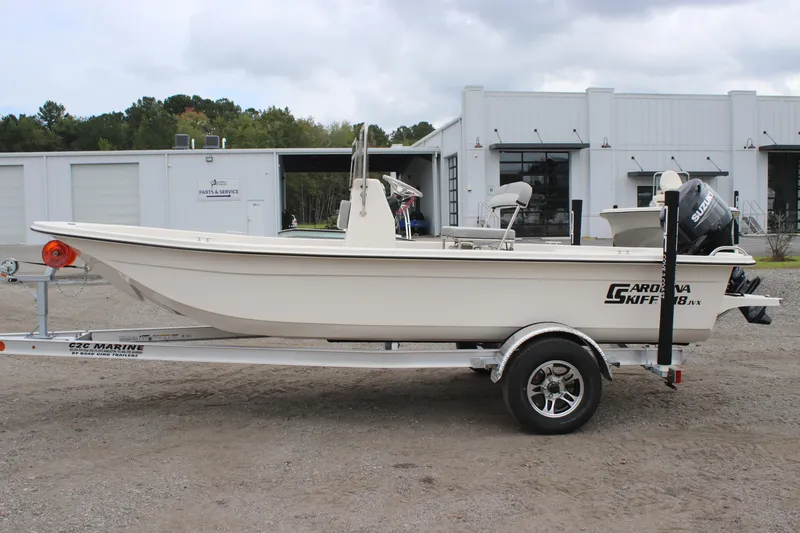 Slide: The Image of 2025 Carolina Skiff E18 JVX CC boat on trailer, parked outdoors near industrial buildings. - 1