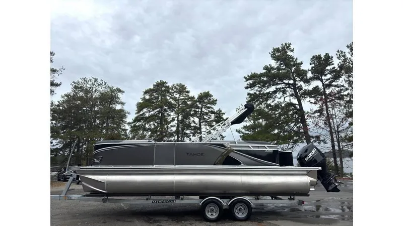 Slide: The Image of 2025 Tahoe Pontoon LTZ Quad Lounger on trailer, parked outdoors with trees in background. - 0