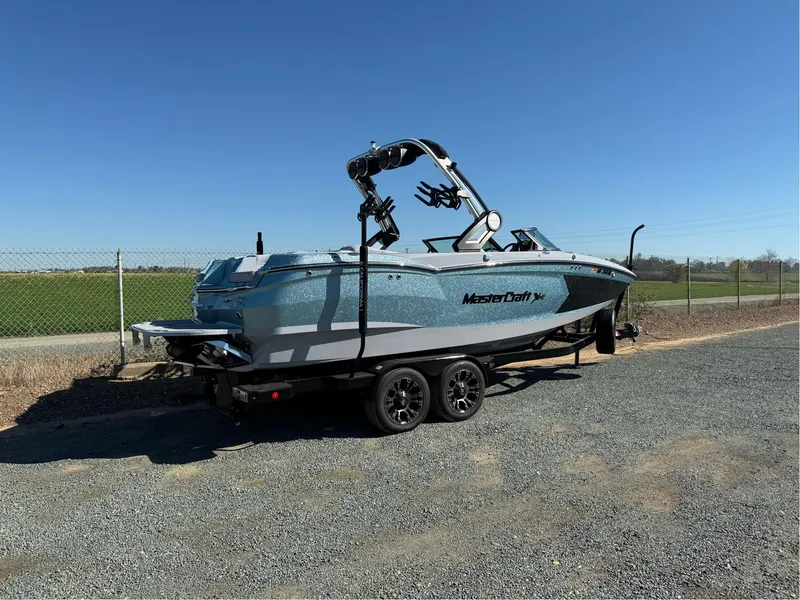 Slide: The Image of 2024 MasterCraft XStar S boat on trailer, parked on gravel near a fence. - 6