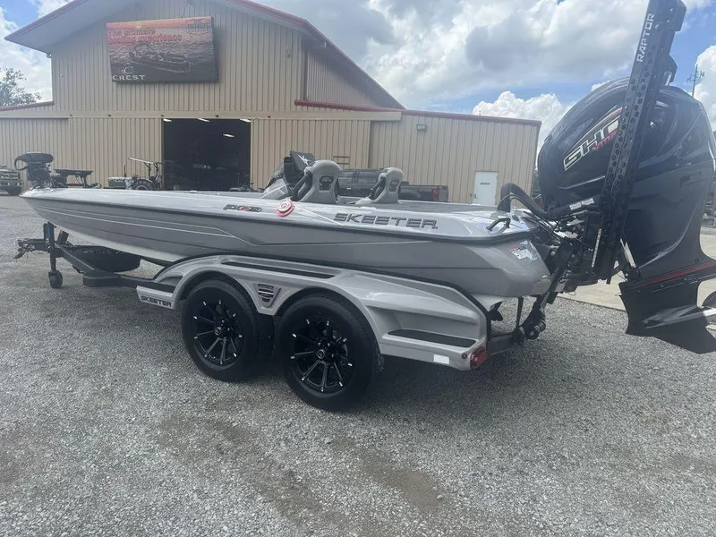 Slide: The Image of 2025 Skeeter FXR20L boat on trailer, parked outside a large building under a cloudy sky. - 2