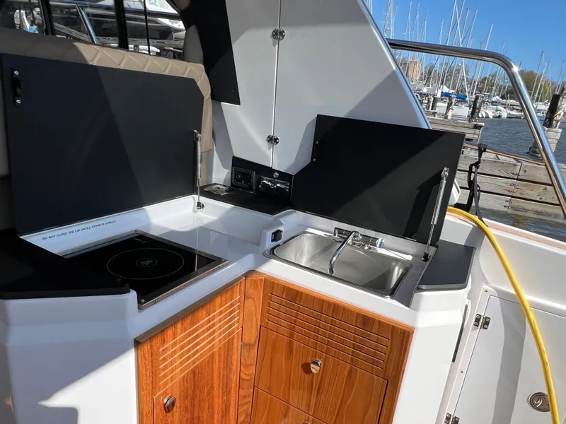 Slide: The Image of 2024 Aquador 250 HT boat kitchen with stove, sink, and wooden cabinetry in a marina setting. - 13