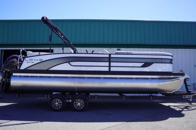 Slide: The Image of 2025 Lowe SS 230 pontoon boat on trailer, parked outside a green building. - 0