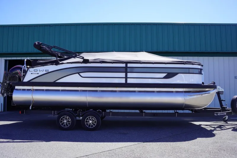 Slide: The Image of 2025 Lowe SS 230 pontoon boat on trailer, parked outside a green building. - 1
