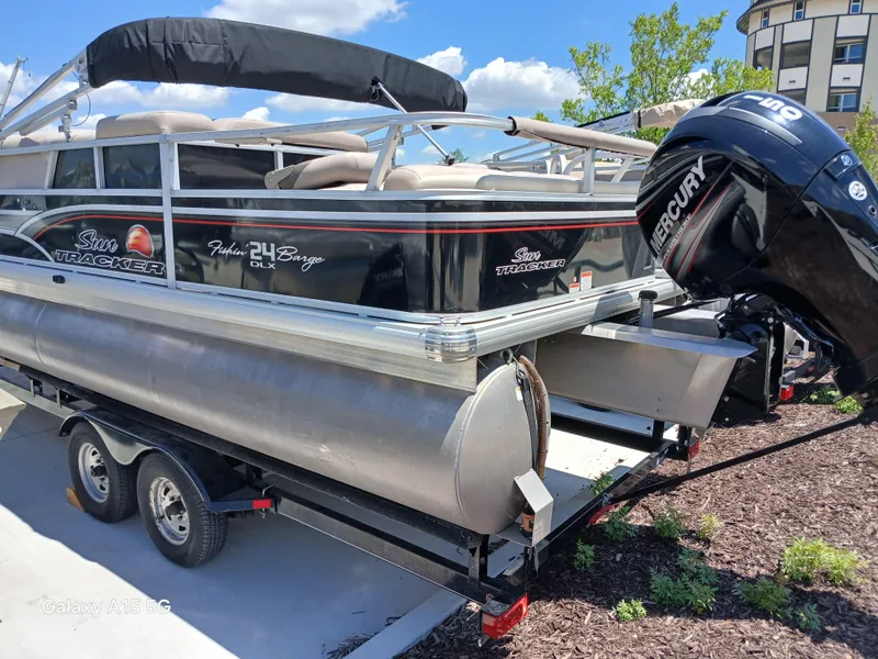 Slide: The Image of 2015 Sun Tracker Fishin Barge 24 DLX pontoon boat on trailer, featuring Mercury outboard motor. - 2