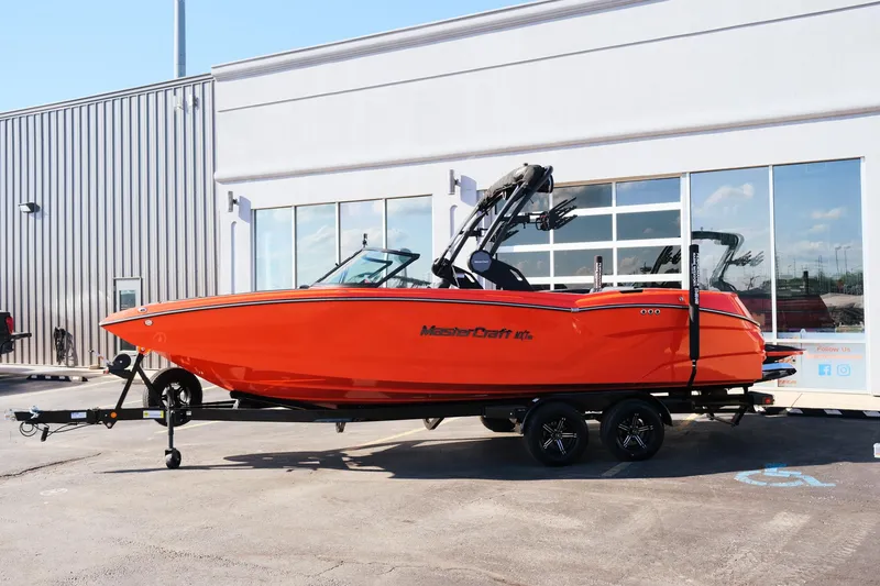 Slide: The Image of 2025 MasterCraft NXT24 boat in vibrant orange, parked outside a modern building. - 2