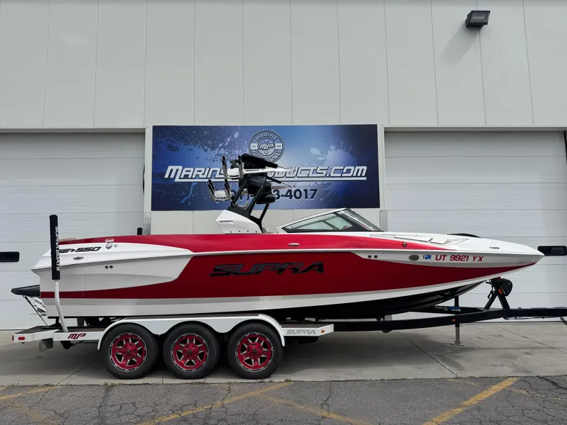Slide: The Image of 2017 Supra SE 550 boat with red and white design on a trailer, parked outside a building. - 0