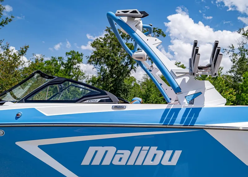 Slide: The Image of 2024 Malibu Wakesetter 24 MXZ boat, blue and white design, sunny day, trees in background. - 12