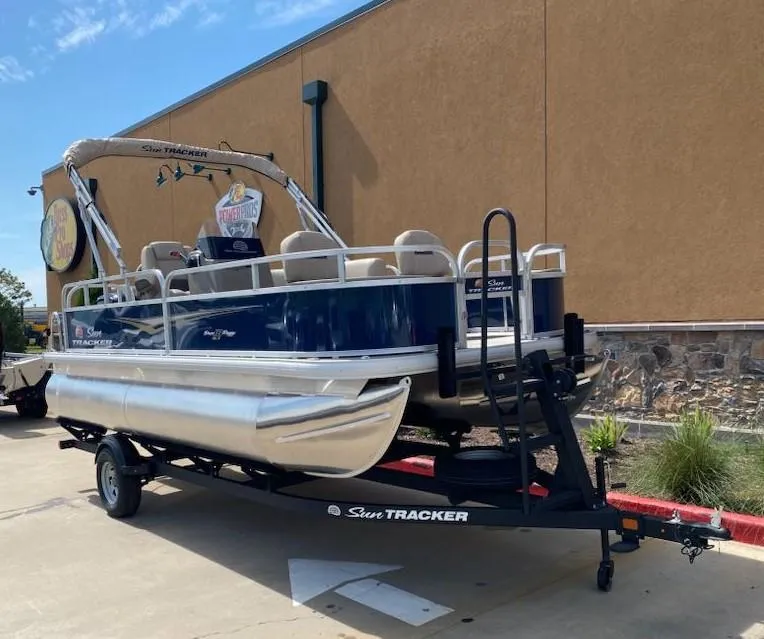 Slide: The Image of 2025 Sun Tracker Bass Buggy 18 DLX pontoon boat on trailer, parked outdoors. - 2