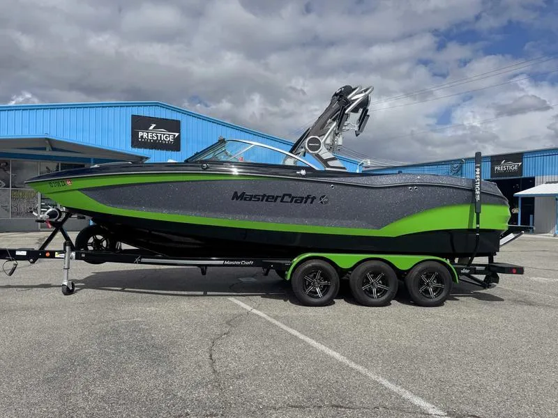 The Image of 2021 MasterCraft X24 boat with green and black design on a trailer, parked outside a blue building. - 1