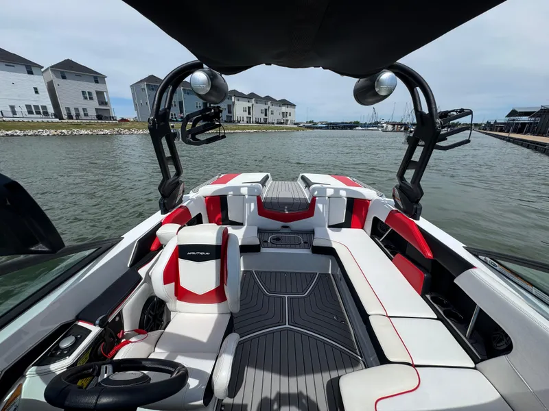 Slide: The Image of 2018 Nautique Super Air Nautique 210 boat interior with red and white seating, docked by waterfront homes. - 13