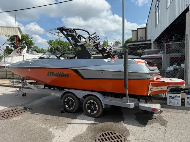 Slide: The Image of 2019 Malibu Wakesetter 22 MXZ boat on trailer, orange and gray design, parked outdoors. - 2