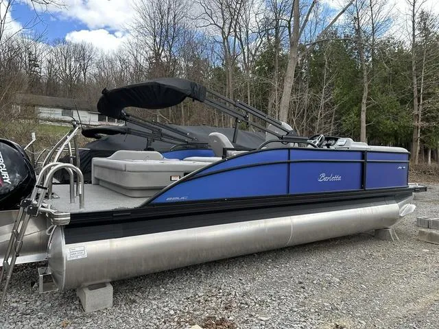 Slide: The Image of 2025 Barletta A22UC pontoon boat with blue exterior, parked on gravel near trees. - 3