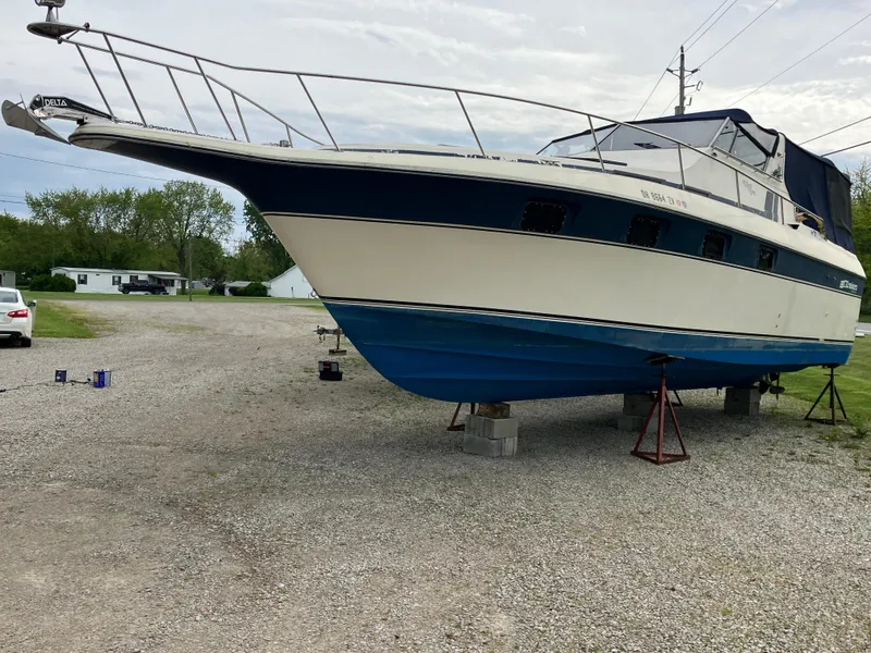Slide: The Image of 1985 Cruisers 336 Ultra Vee boat on stands, parked on gravel, with trees in background. - 5