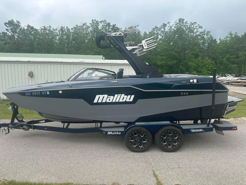 Slide: The Image of 2022 Malibu M220 boat on trailer, parked outdoors near trees and a building. - 24