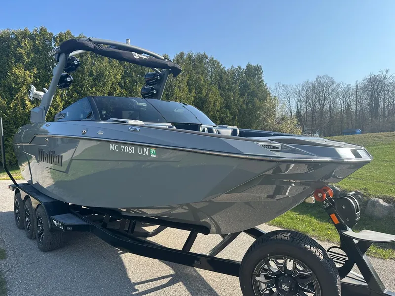 Slide: The Image of 2020 Malibu M240 boat on trailer, parked outdoors, with trees in the background. - 2