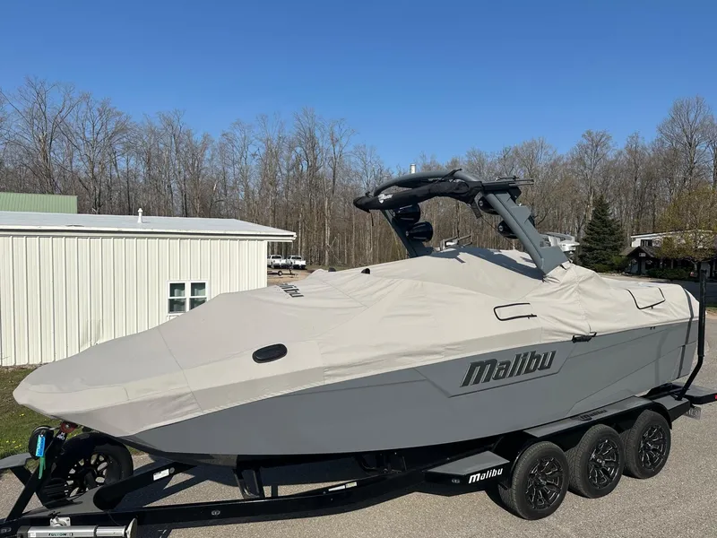 Slide: The Image of 2020 Malibu M240 boat covered on trailer, parked outdoors near trees and building. - 34