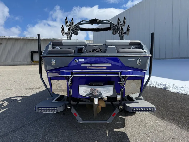 Slide: The Image of 2021 Malibu M240 boat, rear view, blue color, parked outdoors near industrial building. - 2