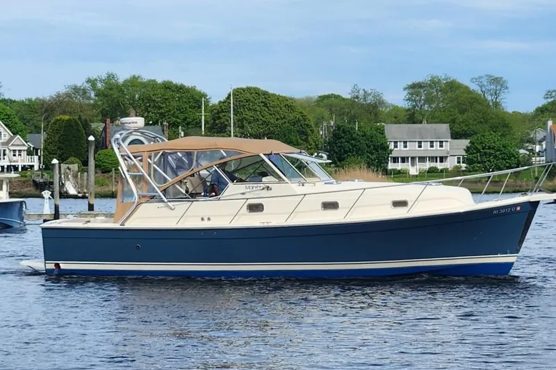 Slide: The Image of 2006 Mainship 34 Rum Runner Classic boat on calm water, with houses in the background. - 0