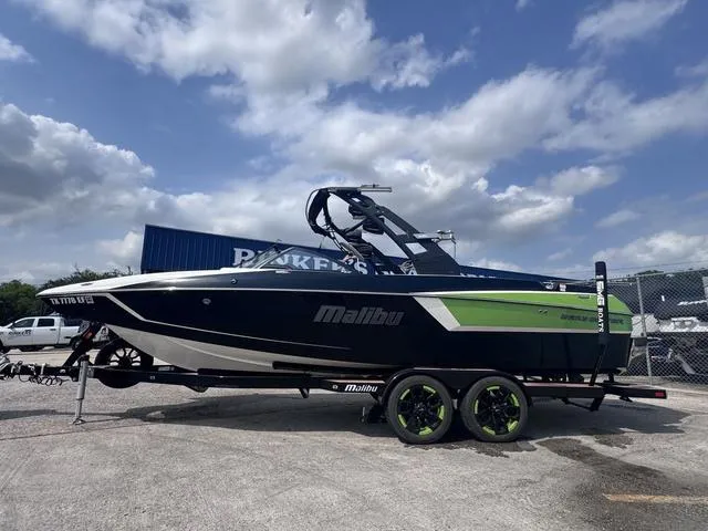 Slide: The Image of 2018 Malibu 24 MXZ boat on trailer, blue and green design, parked outdoors under cloudy sky. - 6