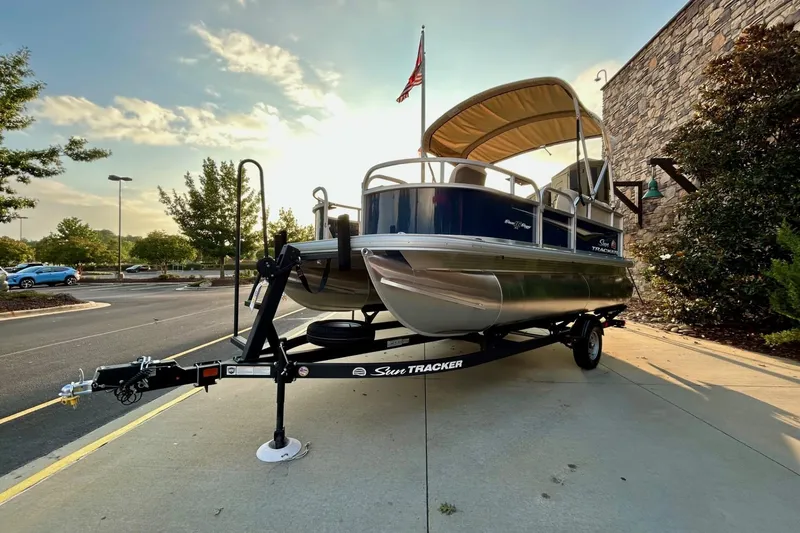 Slide: The Image of 2025 Sun Tracker Bass Buggy 16 XL Select pontoon boat on trailer, parked outdoors. - 2