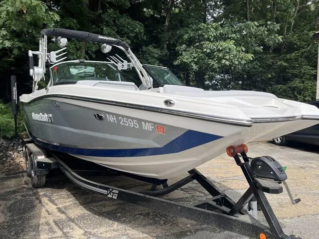 Slide: The Image of 2021 MasterCraft XT22 boat on trailer, parked outdoors with trees in background. - 2
