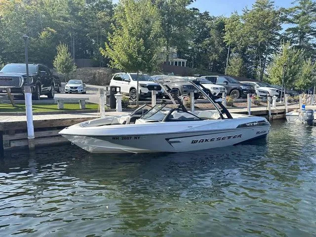 Slide: The Image of 2014 Malibu Wakesetter 23 LSV boat docked by a lakeside with parked cars nearby. - 1